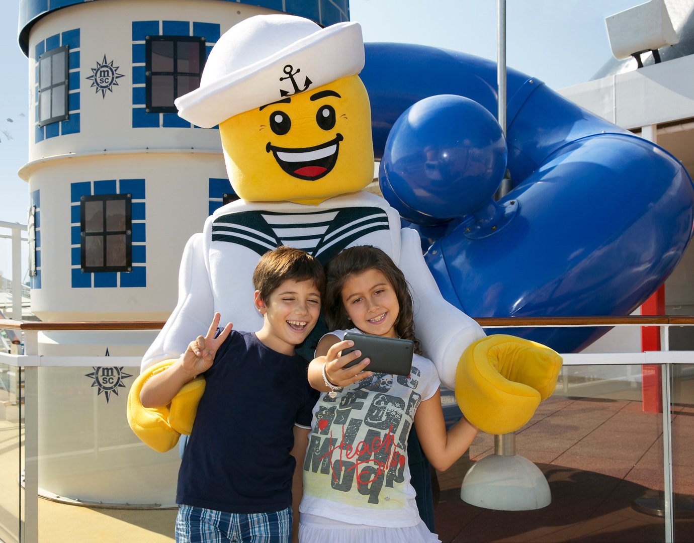 Children taking a selfie with a smiling sailor mascot on an MSC cruise ship deck | MSC Cruises Children taking a selfie with a smiling sailor mascot on an MSC cruise ship deck | MSC Cruises