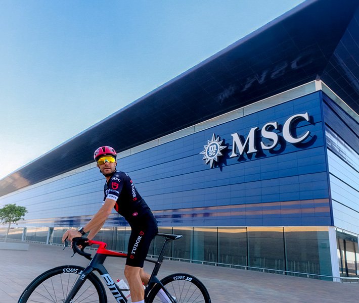 Un ciclista con indumentaria de MSC frente a un edificio moderno de MSC | MSC Cruceros